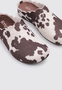 Zuecos de fieltro marrones y crema con estampado de vaca, que presentan un borde ondulado y una textura suave, diseñados para la comodidad y el uso casual.