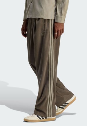 Persona con pantalones de pana marrón sueltos con rayas beige y zapatillas blancas con rayas negras, de pie frente a un fondo claro.