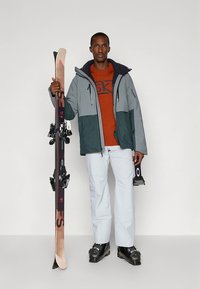 Smučarska jakna v sivi in turkizni barvi, oranžna majica, bele hlače; držanje smuči z vezmi in smučarskimi čevlji. Čiste linije, funkcionalen dizajn.