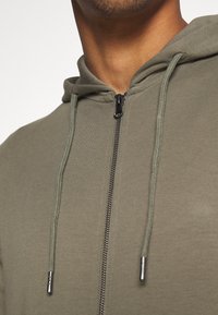 Gros plan d’une personne portant un sweat à capuche zippé vert olive avec des cordons et des embouts métalliques, la fermeture éclair est complètement fermée au niveau du col.