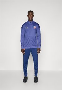 Ljubičasta sportska jakna sa zatvaračem, logo na prsima, uparena s plavim trenirkama. Materijal izgleda lagano, pogodno za sport. Crna obuća.