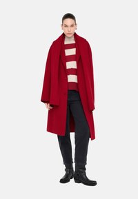 Cappotto di lana rosso con ampi rever, indossato sopra un maglione a righe rosse e crema. Abbinato a jeans neri e stivaletti con dettagli borchiati.