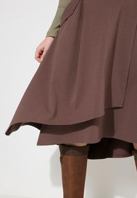 Bruine knielange rok van zacht materiaal, met een flared ontwerp, een gebogen zoom en subtiele plooien. Bruine kniehoogte laarzen zijn zichtbaar.