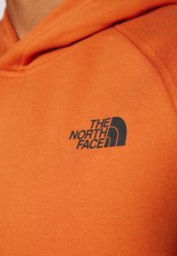 Felpa arancione con una texture morbida, caratterizzata da un logo nero prominente "THE NORTH FACE" sull'area del petto sinistro.