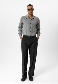 Grijs overhemd met knopen en een gladde textuur, gecombineerd met zwarte broek. Zwarte schoenen met een subtiele glans. Zonnebril maakt de look compleet.