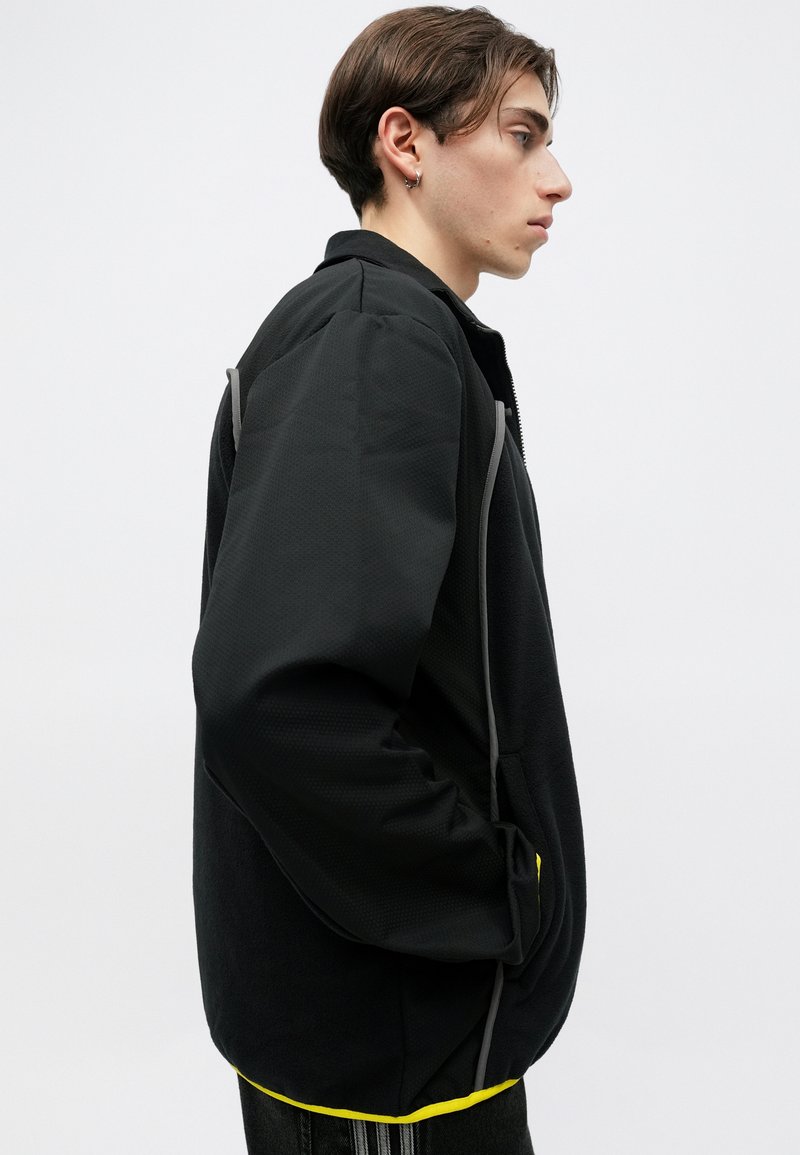 Schwarze Fleecejacke mit strukturiertem Finish, mit Stehkragen, grauen seitlichen Akzenten und gelbem Innenbund. Mit Reißverschlusstaschen vorne.