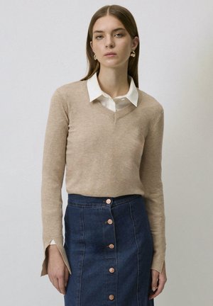 Beige V-hals trui over een witte blouse met kraag, gecombineerd met een donkerdenimrok met koperkleurige knopen en zijsplitten.