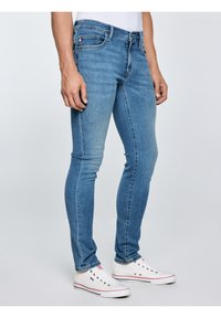 Vyras, dėvintis siaurus mėlynos spalvos džinsus ir baltus žemą kulną turinčius sportbačius, stovintis priešais vientisą šviesią foną.