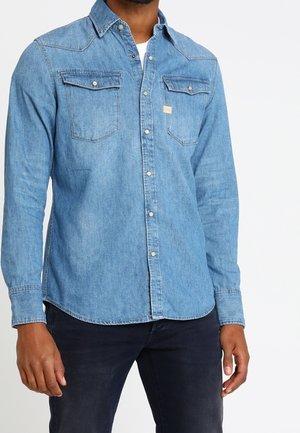 Homme portant une chemise en jean bleu clair à boutons avec poches poitrine et pantalon foncé sur fond uni.