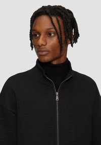 Veste zippée noire en tissu texturé et nervuré, avec un col montant et un détail de fermeture éclair argenté. Col roulé ajusté en dessous.