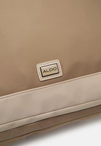 Tan nylon tas met een gladde textuur, met een crèmekleurige accent aan de basis en een geïmpregneerd metalen ALDO-logo.