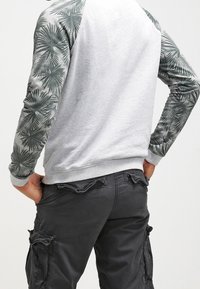 Persona che indossa un felpa grigia chiara con maniche a motivo palme verde scuro e pantaloni cargo grigi scuri, mani nelle tasche posteriori.