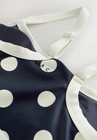 Tissu bleu marine à gros pois blancs avec bretelles blanches réglables, avec un marquage "ACTIVE" partiellement visible.