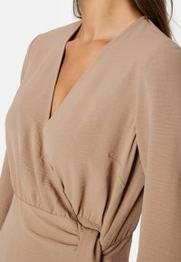 Beżowa sukienka kopertowa z długimi rękawami i głębokim dekoltem w kształcie V. Miękki, lekki materiał z węzłowym detalem w talii. Gładka tekstura.