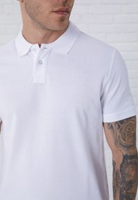 White polo shirt with a classic collar, two-button placket, short sleeves, and a textured fabric. Features a tattoo on the arm.