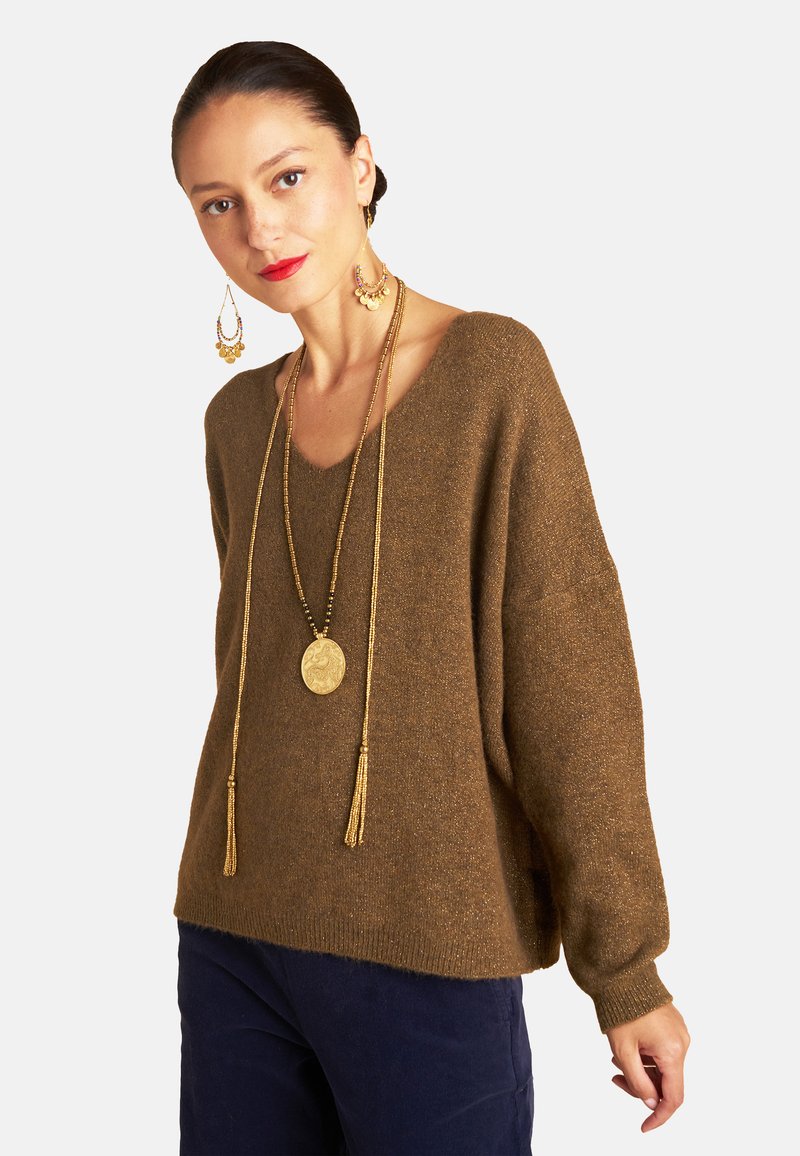 Brauner Strickpullover mit tiefem V-Ausschnitt, weiten Ärmeln und gerippten Bündchen, getragen mit langen goldenen Halsketten und hängenden Ohrringen.