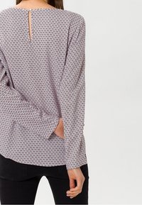 Dlouhý rukáv top vyrobený z lehkého materiálu, který má geometrický vzor v purpurové, červené a bílé barvě; detail na zádech ve tvaru klíčové dírky a uvolněný střih.