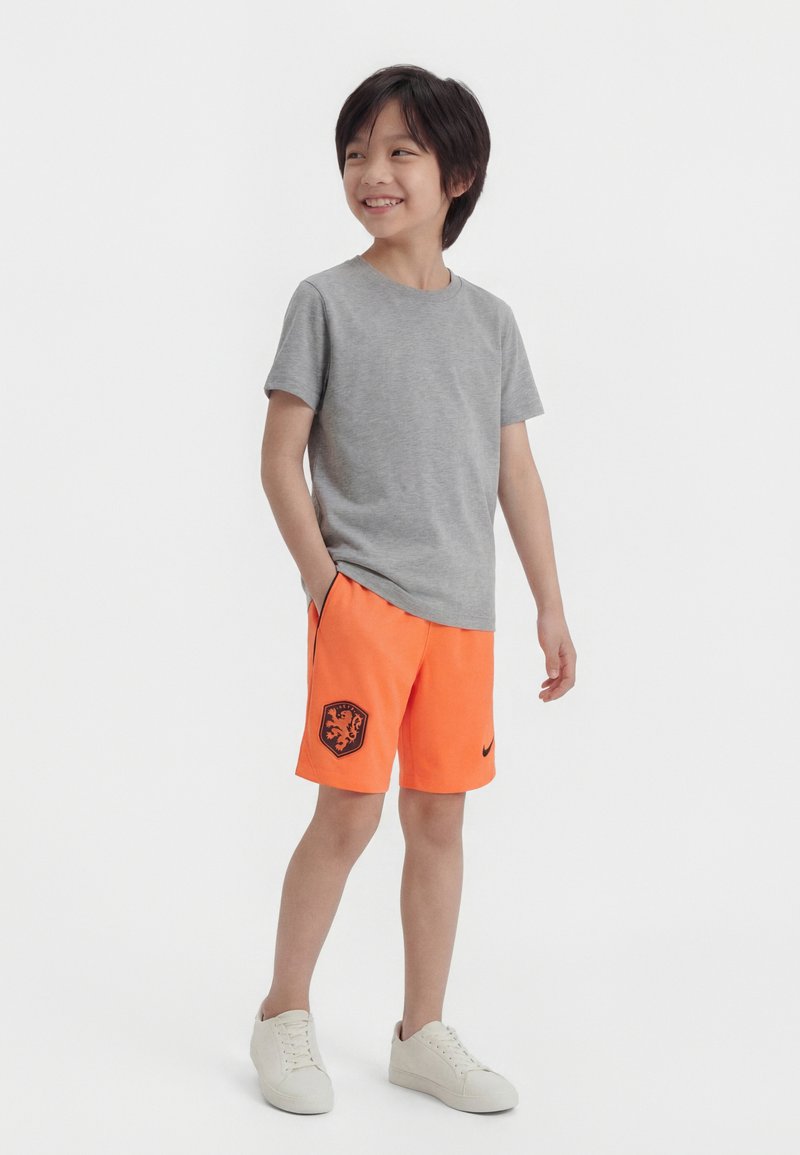 Ένα χαμογελαστό αγόρι στέκεται με τα χέρια στις τσέπες, φορώντας γκρι μπλουζάκι, φωτεινά πορτοκαλί σορτς με μαύρο έμβλημα και λευκά αθλητικά παπούτσια, με φόντο λευκό.