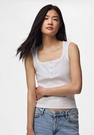 Ung kvinde med langt mørkt hår iført en hvid ærmeløs knapbluse og lyseblå jeans foran en ensfarvet baggrund.