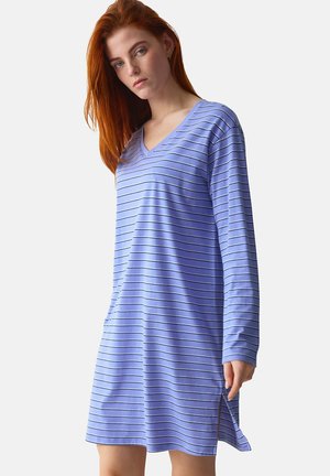 Jonge vrouw met rood haar, gekleed in een blauw-zwart gestreepte V-hals jurk met lange mouwen, staand tegen een eenvoudige lichte achtergrond.