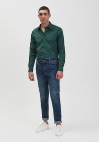 Camicia verde a maniche lunghe con bottoni abbinata a jeans blu e sneakers bianche. La camicia presenta un colletto strutturato e una vestibilità slim.