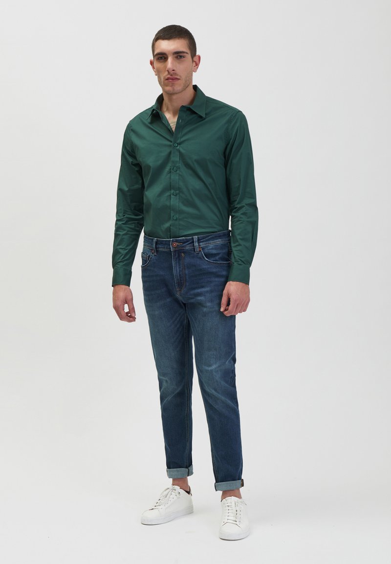 Camicia verde a maniche lunghe con bottoni abbinata a jeans blu e sneakers bianche. La camicia presenta un colletto strutturato e una vestibilità slim.