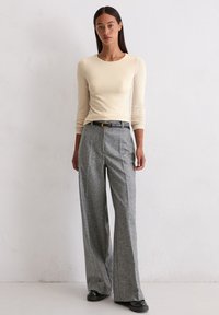 Romantische crème lange mouwen top, gecombineerd met grijze geplooide wijde broek, vastgemaakt met een zwarte riem, aangevuld met zwarte schoenen.