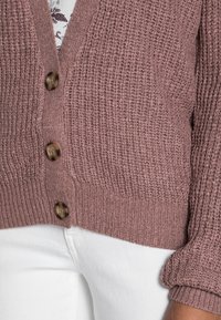 Närbild av en person som bär en lila stickad kofta med bruna knappar över en mönstrad skjorta och vita byxor.