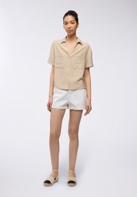 Camicia beige a maniche corte con bottoni e finitura testurizzata, abbinata a pantaloni corti di lino bianchi e scarpe espadrilles chiare con rifiniture nere.