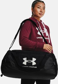 Zwarte sporttas met een groot wit Under Armour-logo, gemaakt van duurzaam materiaal, met dubbele handvatten en een schouderriem voor gemakkelijk dragen.