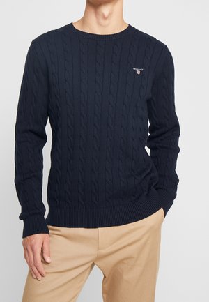 Homme portant un pull en maille torsadée bleu marine avec le logo GANT sur la poitrine et un pantalon beige, debout avec une main dans la poche.