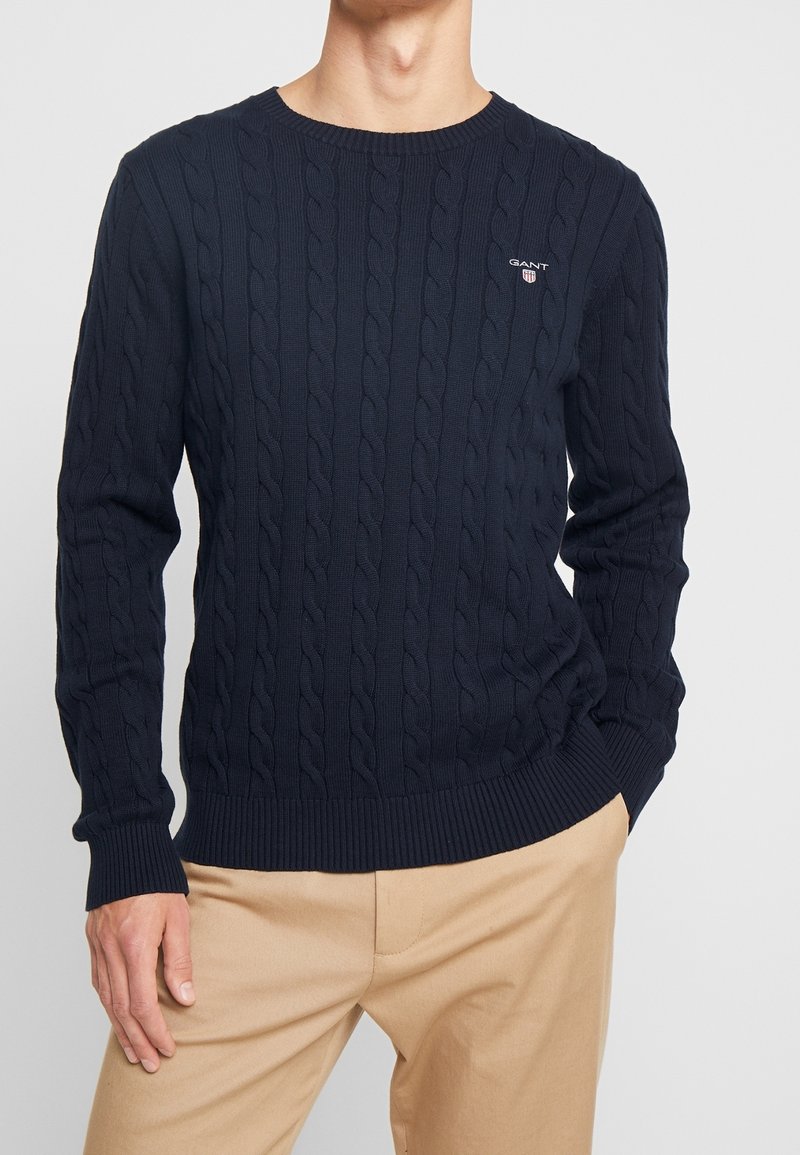 Homme portant un pull en maille torsadée bleu marine avec le logo GANT sur la poitrine et un pantalon beige, debout avec une main dans la poche.