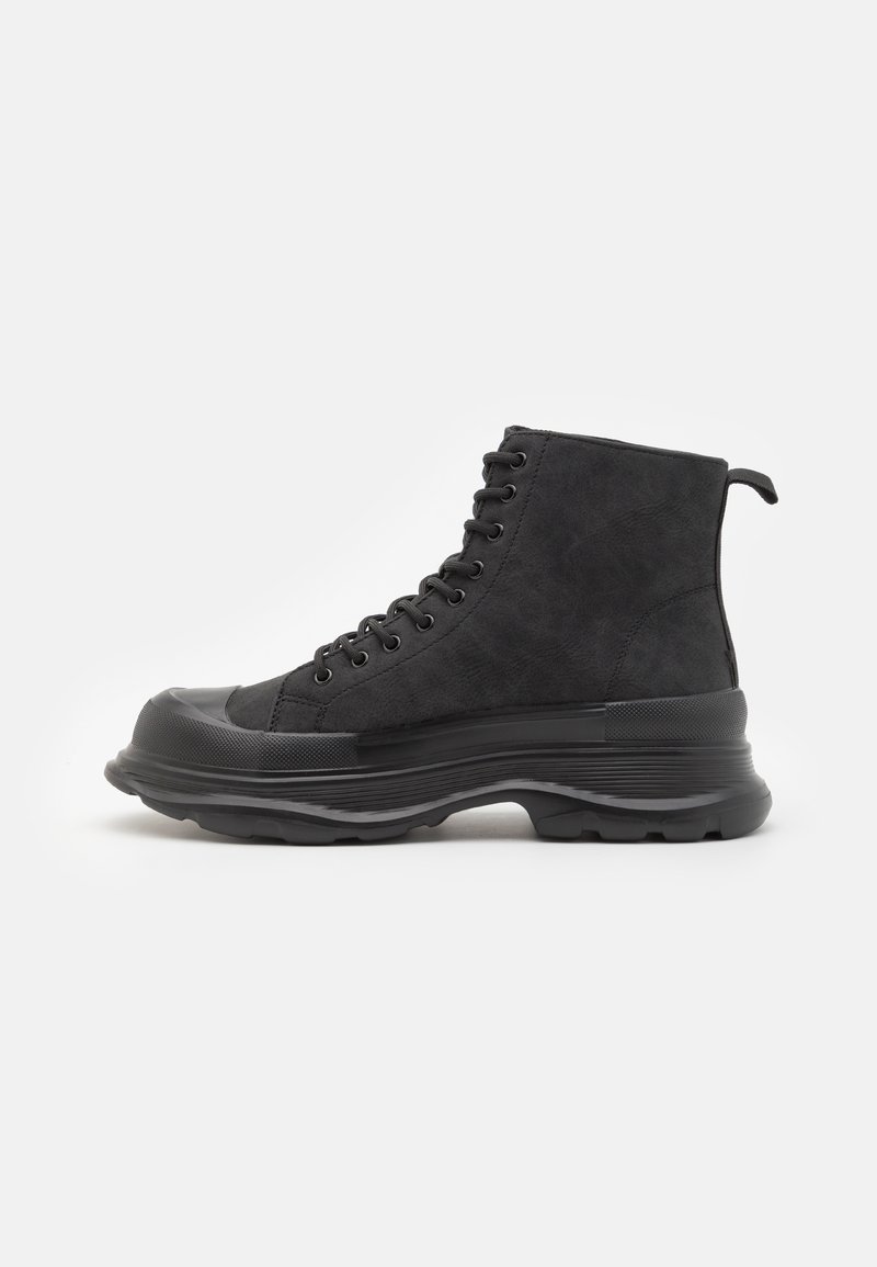 Black ankle boots with a textured upper and rubber toe cap. Features a thick, ridged sole and lace-up design with a pull tab at the back.