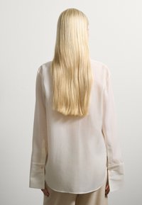 Blusa ligera y transparente hecha de un tejido suave en un beige pálido, con mangas largas y puños amplios, y un dobladillo redondeado.