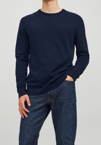 Maglione a maglia blu navy con scollatura rotonda e polsini a costine, abbinato a jeans in denim scuro. Texture morbida, vestibilità standard.