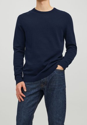Marinblå stickad tröja med rund halsringning och ribbade muddar, tillsammans med mörka jeans. Mjuk textur, normal passform.