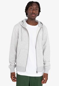 Šedá zipová mikina z bavlnenej zmesi, s kapucňou na šnúrku, prednými vreckami a rebrovanými manžetami. Nosená cez biele tričko a zelené nohavice.