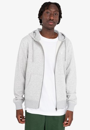 Grå zip-up hoodie med en bomuldsblanding, der har en snøre i hætten, forkert lommer og ribbede manchetter. Bæres over en hvid t-shirt og grønne bukser.