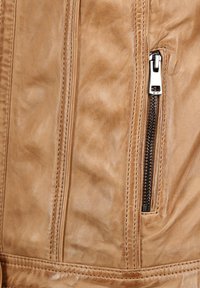 Detail van een bruine leren jas met een diagonale ritszak met zilverkleurige hardware, een gladde textuur en contrasterende stiksels langs de naden.