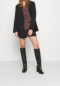 Blazer noir sur un chemisier à motifs avec des accents rouges, associé à une mini-jupe noire et des bottes noires à talons montantes jusqu'aux genoux.