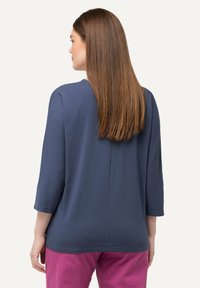 Blaues Oberteil mit Dreiviertelärmeln, rundem Ausschnitt und lässiger Passform. Die Rückseite hat eine zentrale Naht und wird mit rosa Hosen kombiniert.