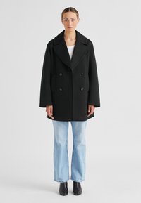 Schwarzer doppelt knopfbarer Mantel mit breitem Revers, Seitentaschen und lässiger Passform, kombiniert mit hellblauen, weit geschnittenen Jeans und schwarzen Stiefeln.