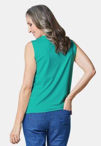 Mouwloze teal top van gladde stof, met een ronde halslijn en een relaxte pasvorm. Gekozen in combinatie met blauwe denim jeans, met zicht op de achterkant.