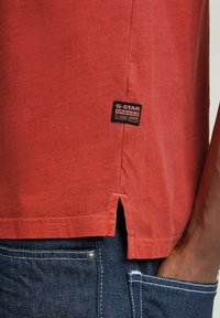 T-shirt in cotone rosso con orlo arrotondato e slits laterali; etichetta attaccata con il nome del marchio "G-Star Originals." Abbinato a jeans in denim scuro.