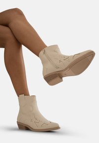 Botines beige hasta el tobillo con diseños florales calados, material de cuero suave, tacón bajo de tipo ondero y detalle de cremallera lateral. Suela de goma para mayor tracción.