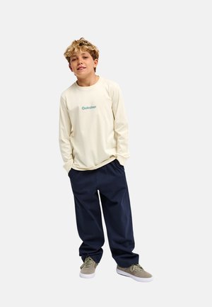 Chłopiec o blond włosach, ubrany w kremową koszulkę Quiksilver z długim rękawem, ciemnoniebieskie spodnie i beżowe trampki, stoi z rękami w kieszeniach.