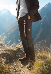 Personne portant un pantalon noir The North Face et des chaussures de randonnée, debout sur un sentier de montagne aux bords herbeux et aux sommets lointains en plein jour.