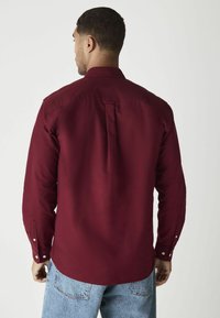 Burgundská košeľa s dlhým rukávom vyrobená z textúrovanej látky s detailom na zadnom diely a gombíkovými manžetami. V kombinácii s svetlomodrými džínami.