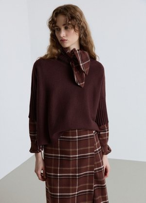 Donna con capelli mossi che indossa un maglione viola scuro sopra un vestito marrone a quadri con una cravatta abbinata, in piedi davanti a uno sfondo semplice.
