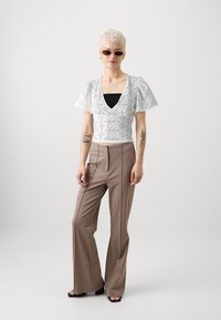 Weißes Crop-Top mit einem Rautenmuster, Puffärmeln über einem schwarzen Tanktop. Braune, ausgestellte Hose mit glatter Textur und minimaler Detailierung. Dunkle Sonnenbrille.